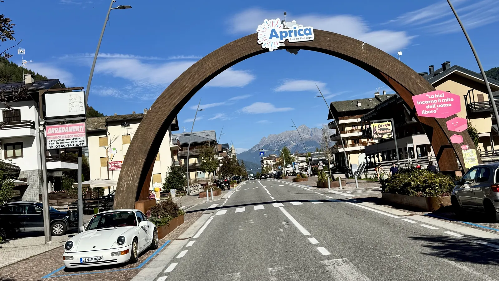 Weißer Porsche 911 unter dem Holzbogen am Ortseingang von Aprica