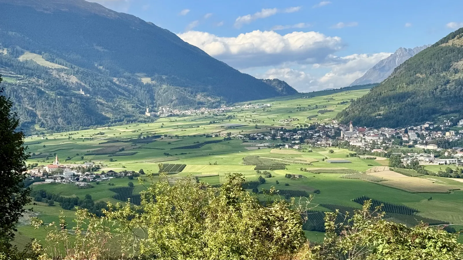 Weites grünes Alpental mit Ortschaften und Bergen unter blauem Himmel