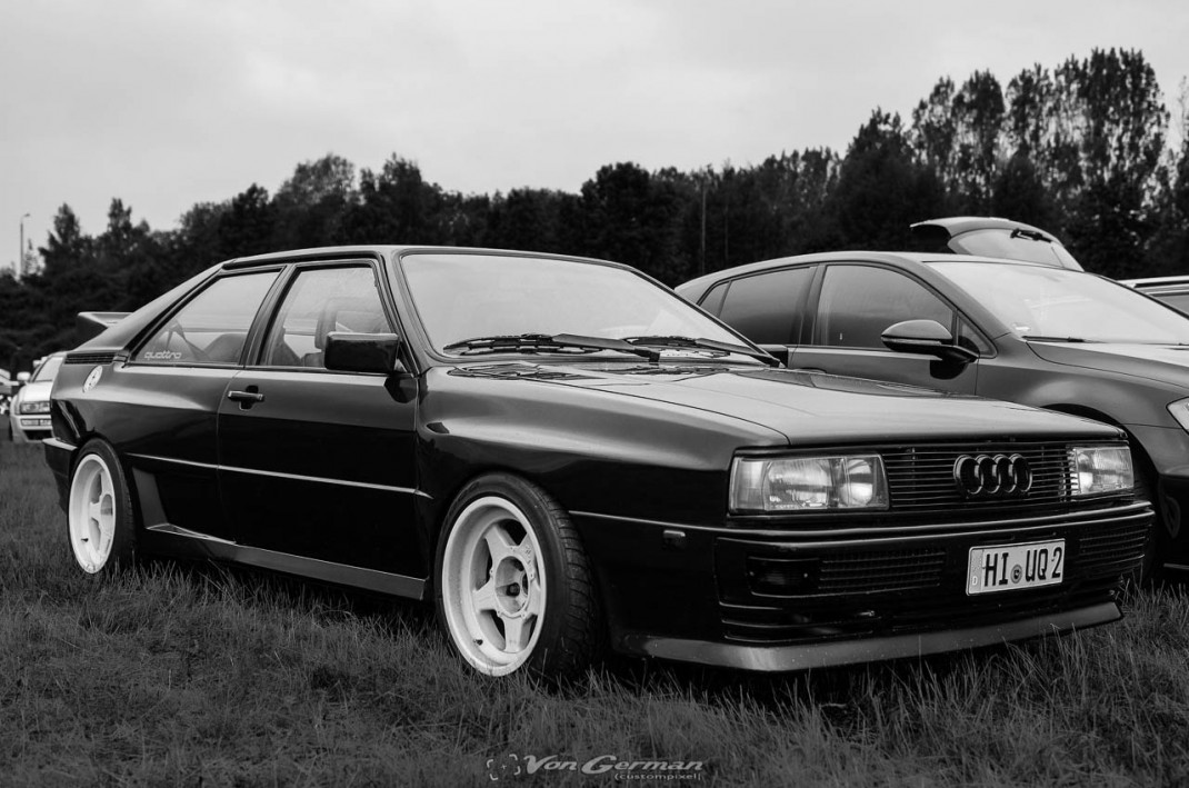 Schwarzer Audi-Oldtimer mit weißen Felgen auf Wiese beim Audi-Treffen, im Hintergrund Nadelwald und weitere Fahrzeuge