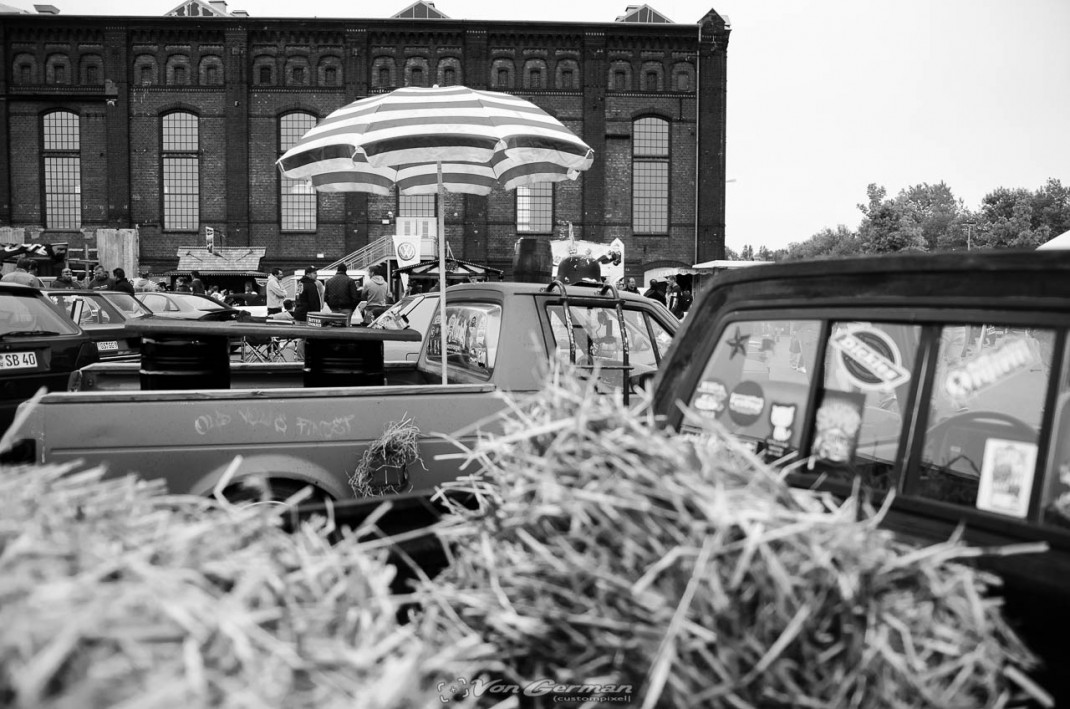 Oldtimer und Youngtimer auf Parkplatz vor Backsteingebäude, Besucher unter Sonnenschirm beim VW & Audi Treffen