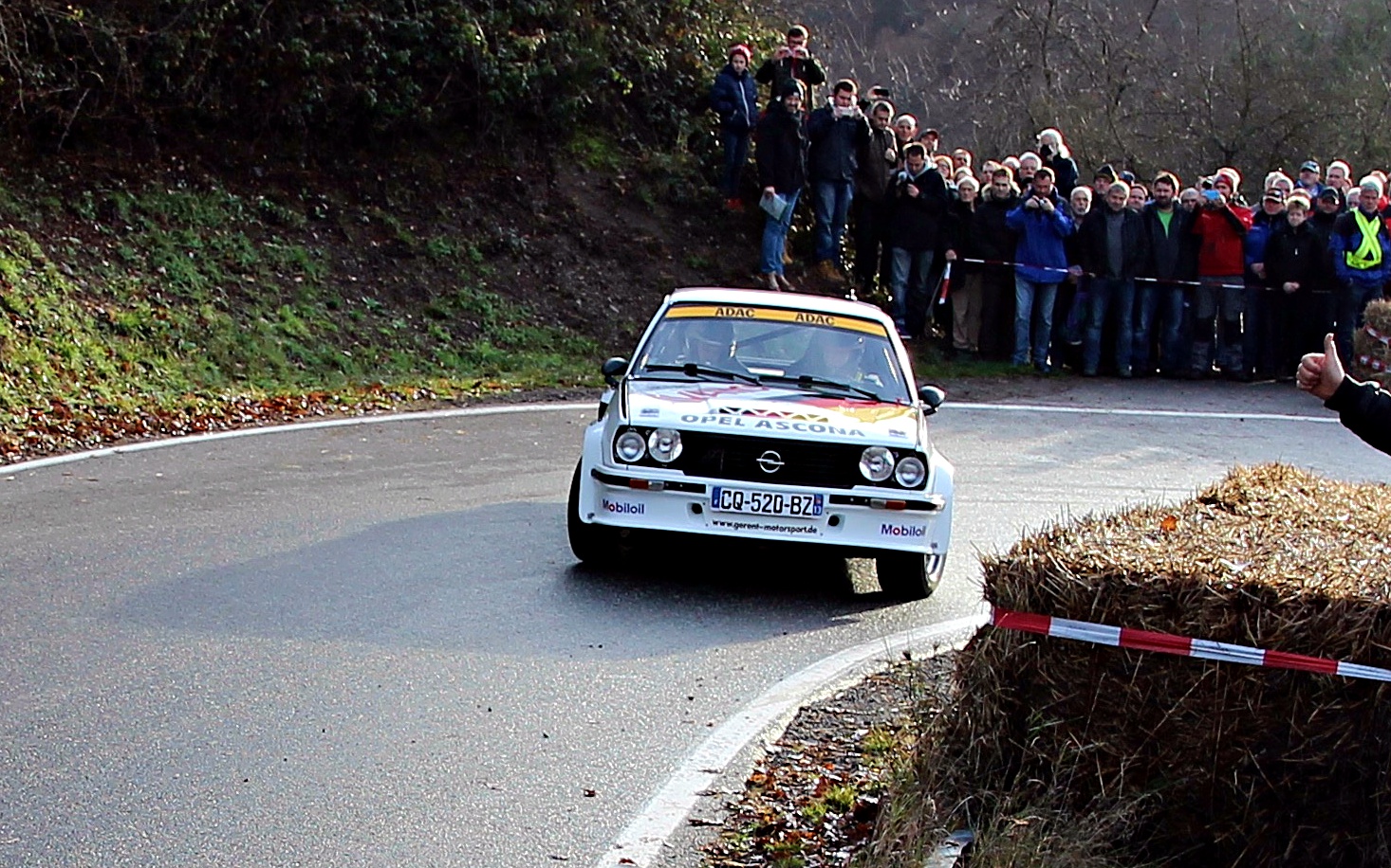 Youngtimer-Rallye-Auto mit vier Scheinwerfern und Startnummer auf kurvenreicher Waldstraße, Zuschauer säumen die Strecke