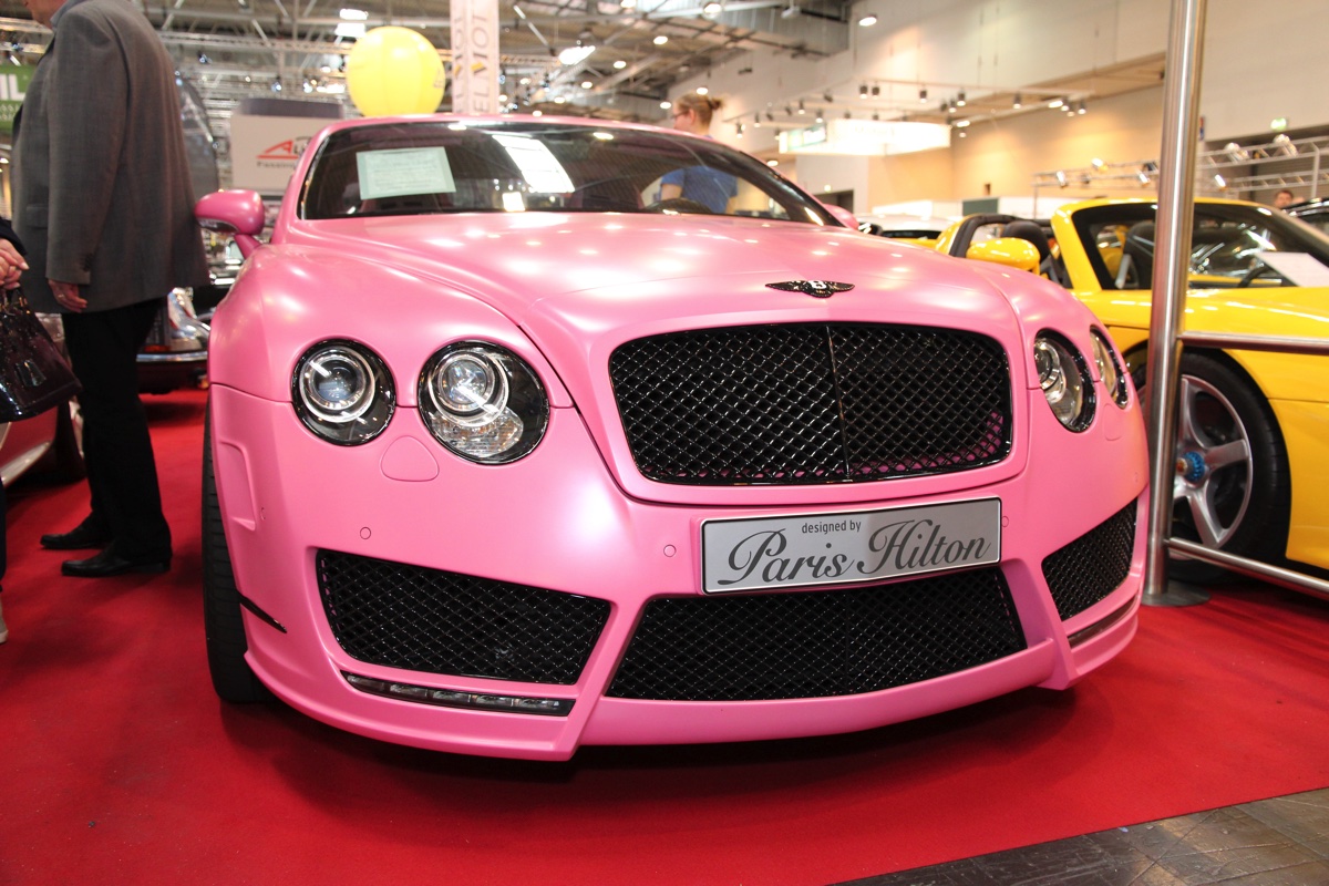 Pink lackierter Luxus-Sportwagen auf rotem Teppich in einer Messehalle, daneben Besucher und weitere Fahrzeuge