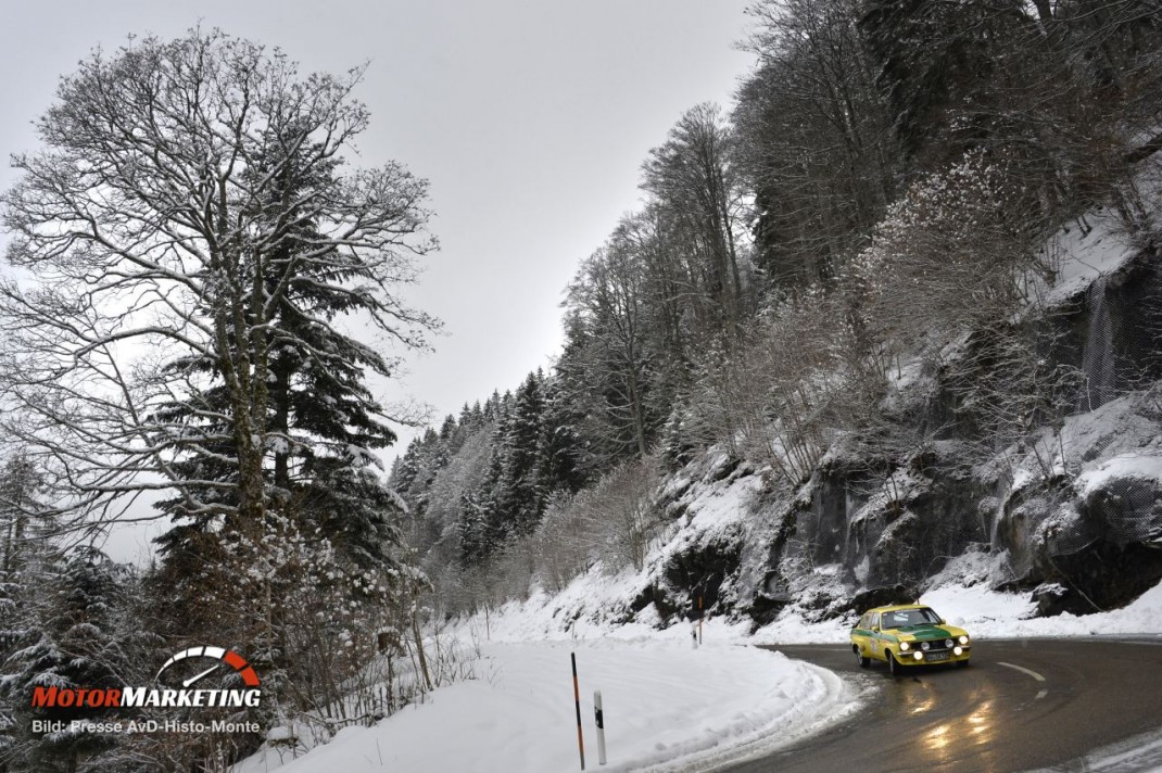 Gelber Youngtimer auf schneebedeckter Bergstraße mit Felswand und Wald in Winterszenerie