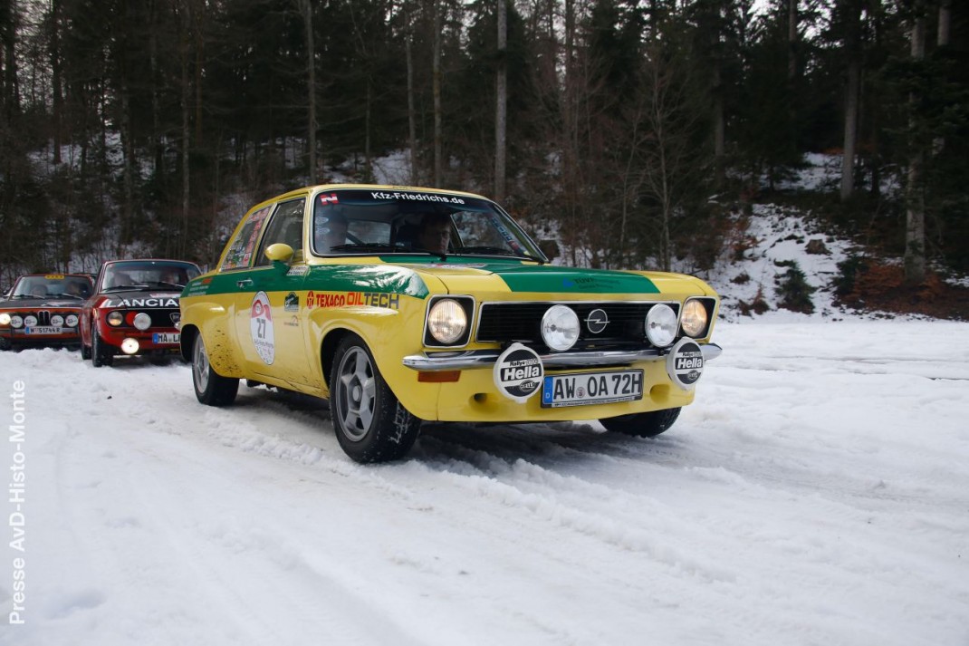 Gelber Opel Ascona mit Rallye-Beschriftung und Zusatzscheinwerfern auf verschneiter Strecke, begleitet von weiteren Oldtimern bei der AvD-Histo-Monte