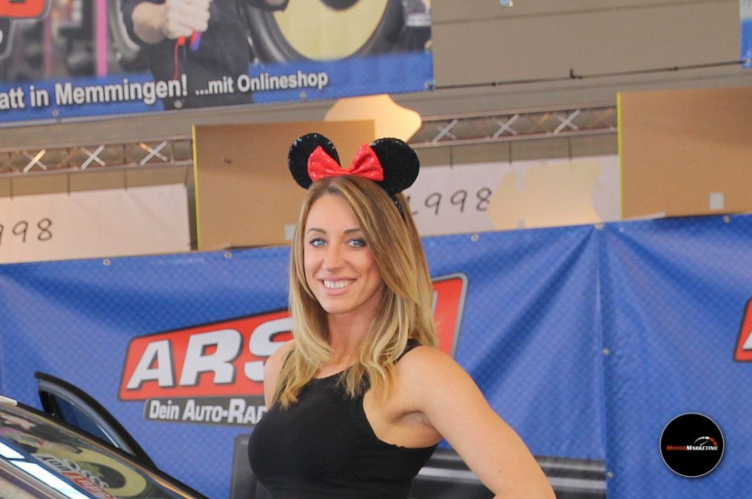 Junge Frau mit Maus-Ohren-Haarreif posiert bei der TuningWorld Bodensee vor einem Auto-Reparatur-Messestand