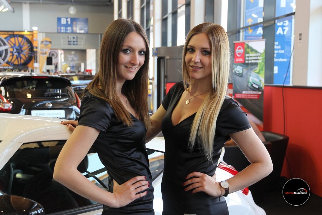 Zwei Moderatorinnen in schwarzen Tops posieren neben einem weißen Sportwagen auf der TuningWorld Bodensee