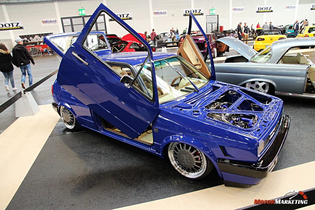 Blauer Youngtimer mit geöffneter Motorhaube und Flügeltüren auf der Automesse, dahinter weitere Oldtimer und Besucher