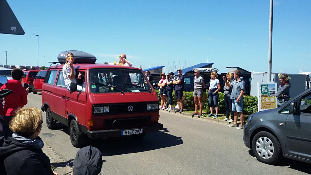 Roter VW-Bulli mit Dachgepäck fährt über Festivalgelände, Zuschauer und andere Fahrzeuge ringsum