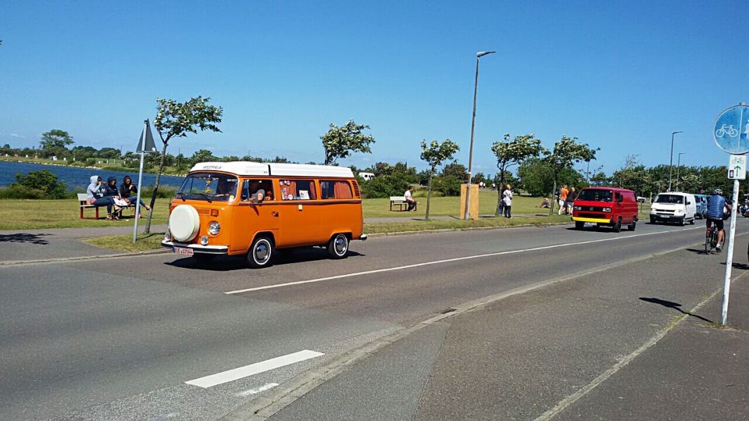 Orangefarbener VW-Bus mit Ersatzrad fährt über Parkplatz beim Bulli-Festival auf Fehmarn, im Hintergrund weitere Oldtimer und Teilnehmer