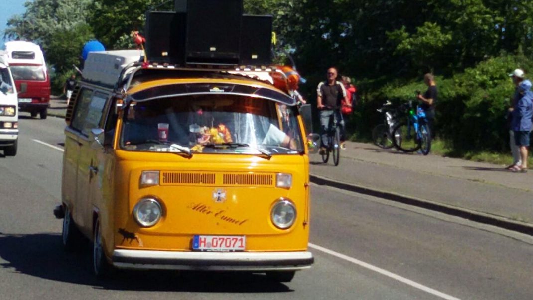 Gelber VW-Bus mit Dachgepäck fährt beim Midsummer Bulli Festival auf Fehmarn, begleitet von Zuschauern und anderen Fahrzeugen am Straßenrand