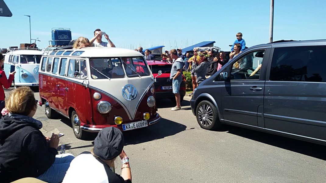 Rot-weißer VW Bulli zwischen anderen Fahrzeugen auf dem Midsummer Bulli Festival auf Fehmarn, Besucher fotografieren und schauen sich die Oldtimer an