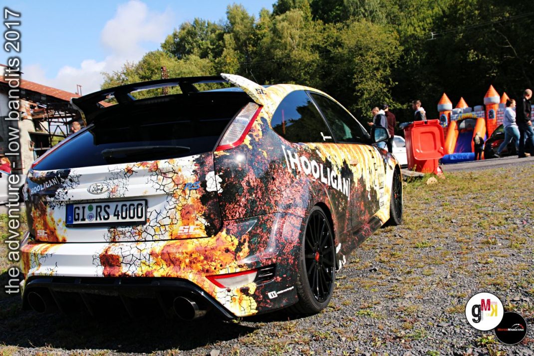 Youngtimer mit Rust-Look-Folierung und offener Heckklappe auf Tuning-Event im Steinbruch, Besucher im Hintergrund