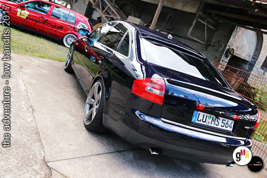 Youngtimer-Limousine in Schwarz-Weiß-Rot auf Parkplatz, im Hintergrund roter Oldtimer und Industriegebäude