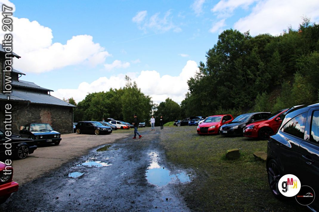 Parkplatz mit Oldtimern und Youngtimern auf Wiese, Steingebäude links, Besucher zwischen roten und blauen Fahrzeugen
