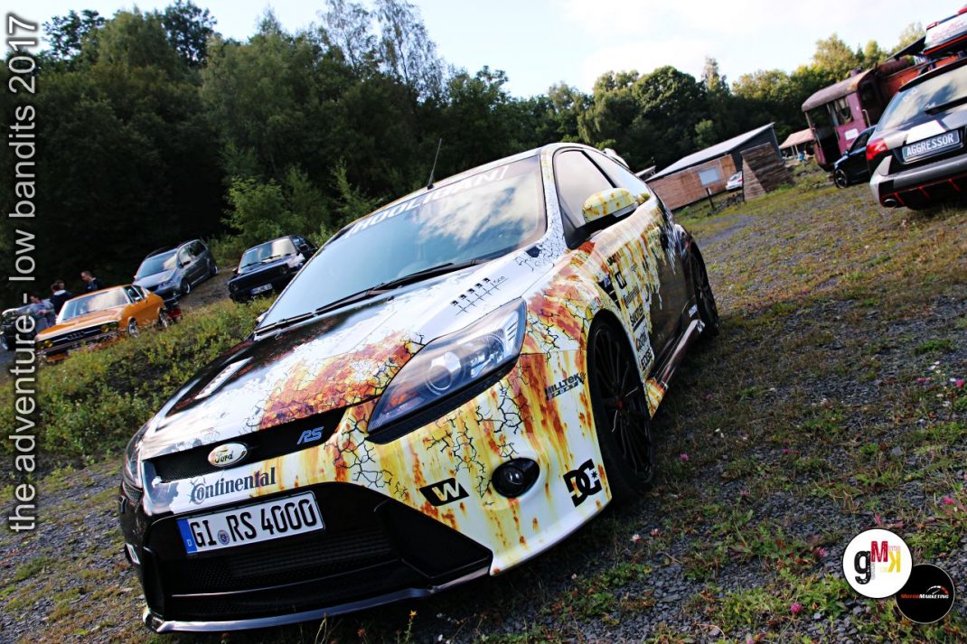 Youngtimer-Sportlimousine mit künstlerischer Rostoptik-Folierung auf Veranstaltungsgelände mit Wald und weiteren Fahrzeugen im Hintergrund