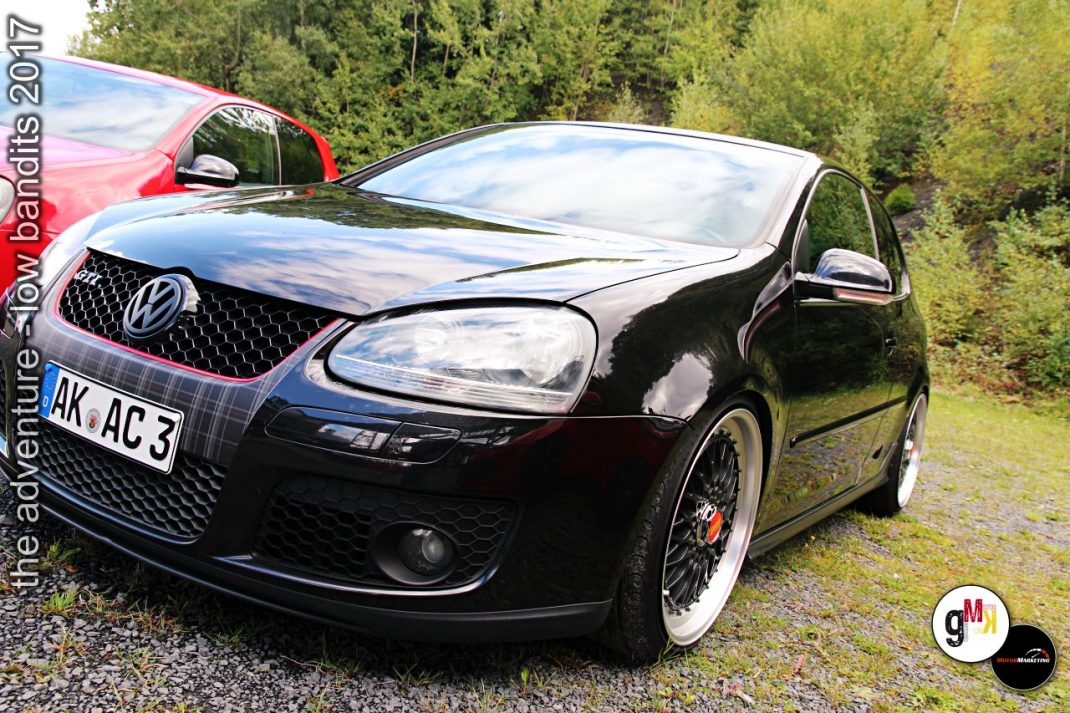 Schwarzer VW Golf mit Tieferlegung und roten Felgen vor grünem Waldgrund, dahinter rotes Cabrio bei Tuning-Event