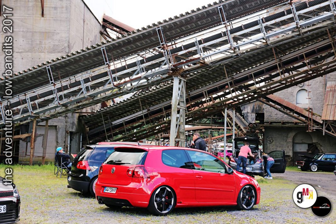 Roter Youngtimer-Kompaktwagen mit schwarzem Dach auf Event-Gelände unter Industriebrücke, Teilnehmer im Hintergrund