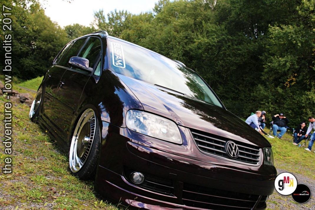 Schwarzer VW Touran mit tiefergelegtem Fahrwerk und Chromfelgen auf Tuning-Veranstaltung im Grünen, Zuschauer im Hintergrund