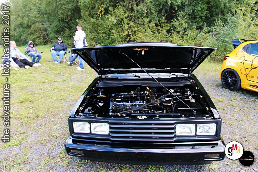 Schwarzer Youngtimer mit offener Motorhaube auf Treffen, Zuschauer im Grünen, gelber Sportwagen rechts