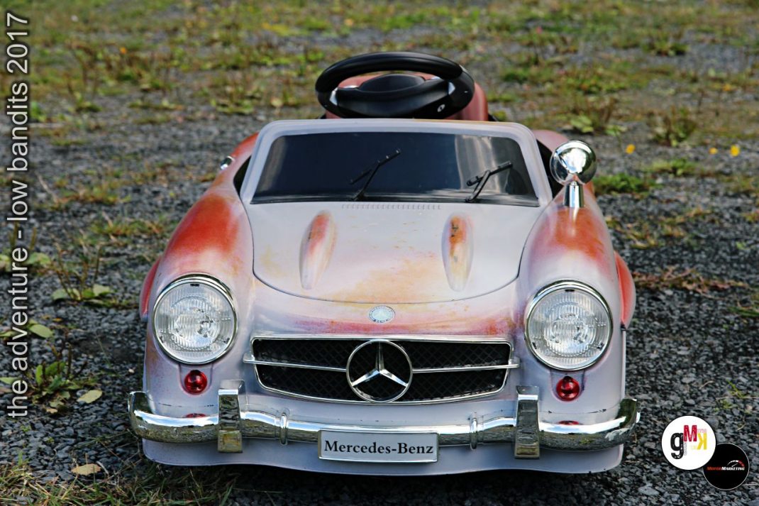 Oldtimer-Kinderauto Mercedes-Benz in Rot-Weiß auf moosigem Untergrund, Frontansicht mit charakteristischem Stern-Emblem
