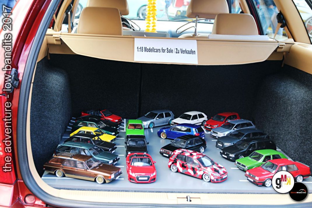 Modellautos verschiedener Farben und Stile im offenen Kofferraum eines roten Oldtimer-Cabrios, Schild „1:18 Modelcars for Sale Zu Verkaufen