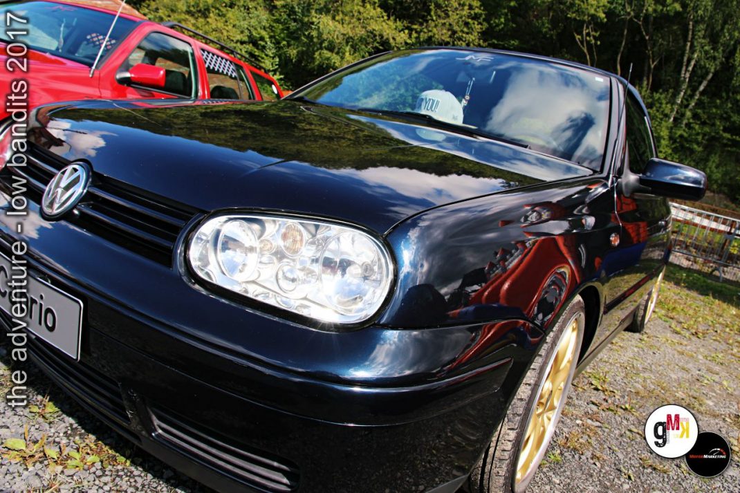 Schwarzer VW Golf Youngtimer mit roten Accents und roten Felgen auf Veranstaltungsparkplatz, im Hintergrund rotes Fahrzeug und Grünfläche
