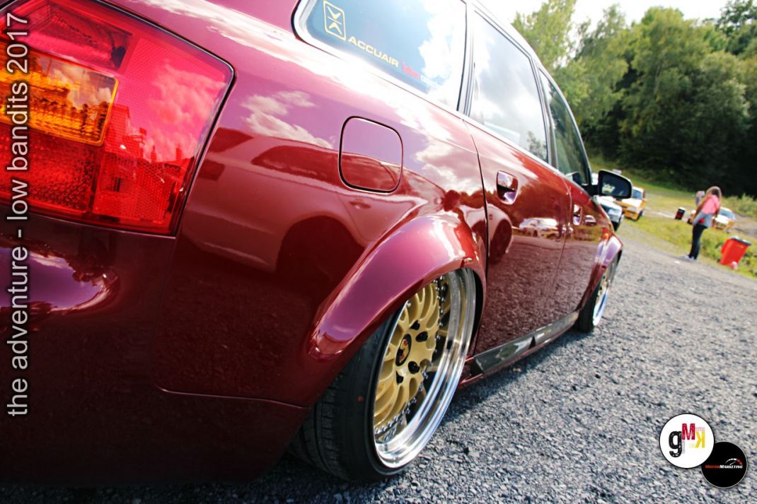 Roter Youngtimer mit goldenen Leichtmetallrädern auf Rallye-Parkplatz, im Hintergrund Teilnehmer und Bäume