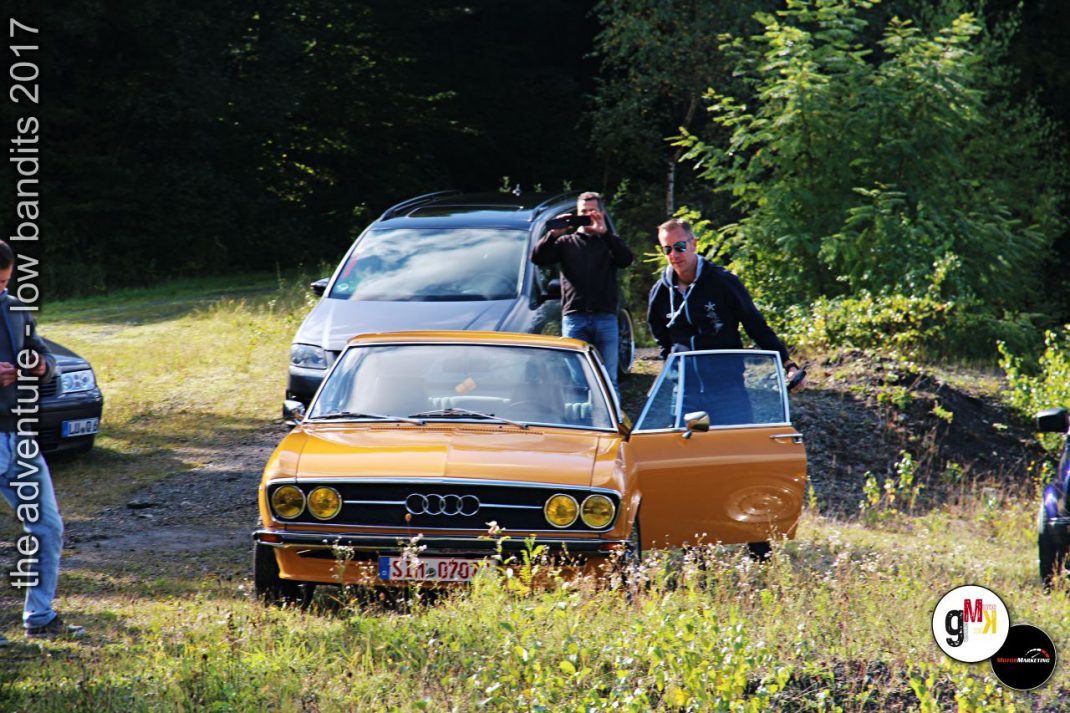Orangefarbener Audi-Oldtimer mit offener Tür und Teilnehmern bei der Low-Bandits-Veranstaltung 2017 in waldiger Umgebung