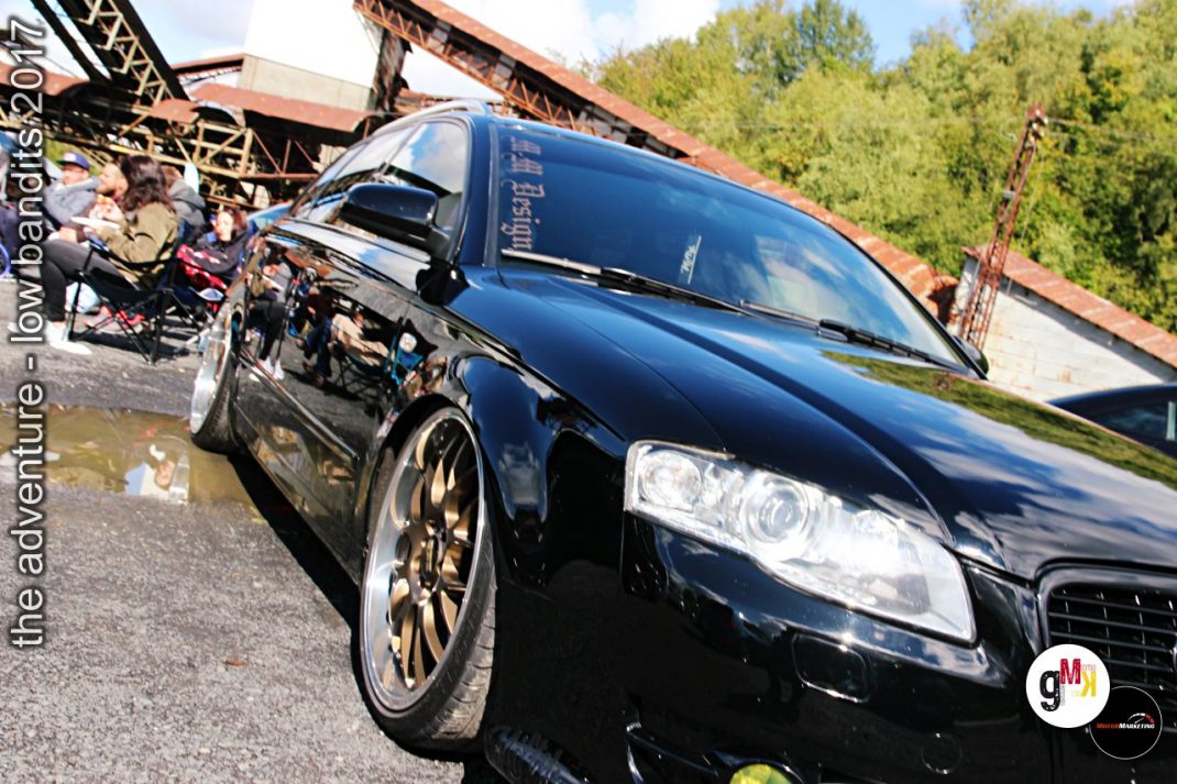 Schwarzer Youngtimer-Limousine mit Sportfelgen auf Gelände mit rostigen Industriebauten und Zuschauern bei Tuning-Event