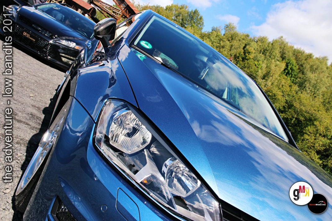 Blaue Youngtimer-Limousine im Detail mit Scheinwerfer und Kühlergrill, daneben weiterer Wagen auf Parkplatz mit Bäumen