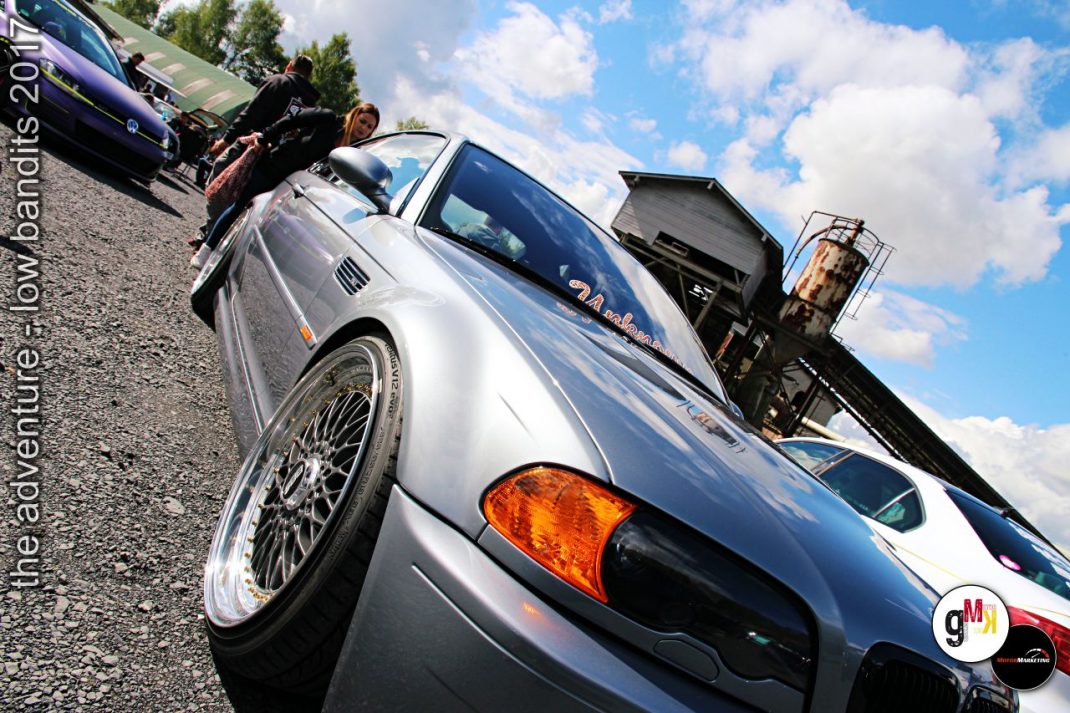 Silberner Youngtimer-Cabrio mit Sportfelgen und schwarzem Hardtop auf Parkplatz bei Tuning-Event, weitere Fahrzeuge im Hintergrund