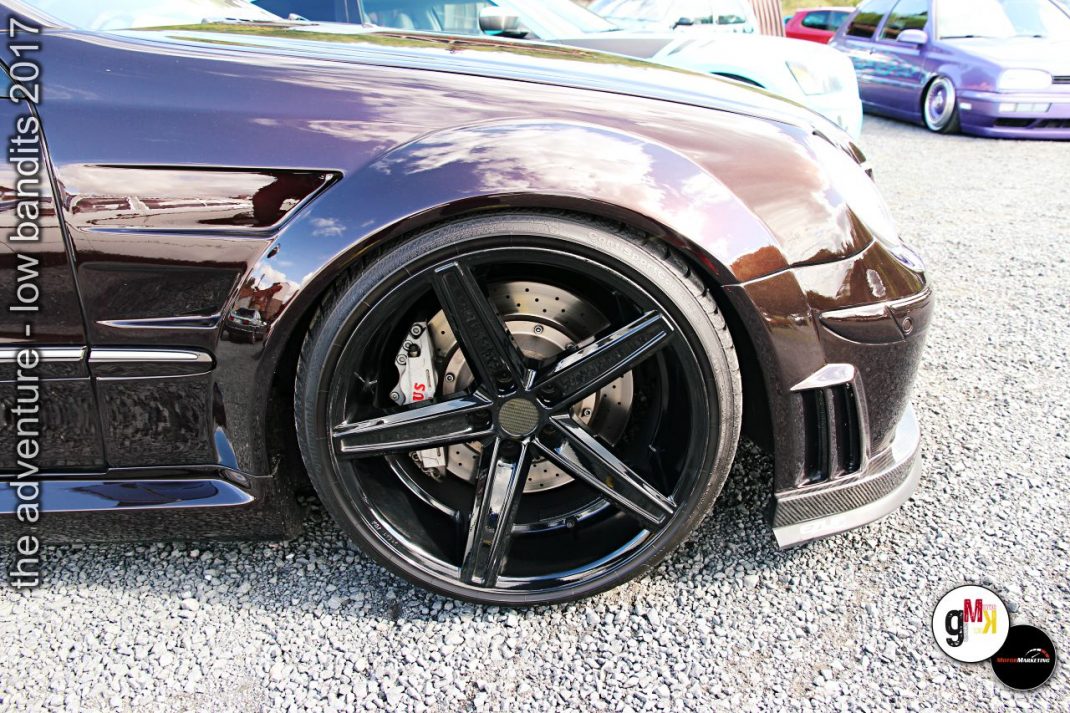 Detailaufnahme eines tiefergelegten Sportwagens mit schwarzem Leichtmetallrad und roten Bremssätteln auf Kiesplatz bei Tuning-Treffen
