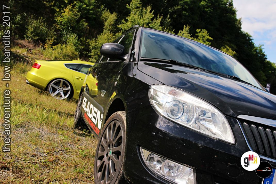 Gelber Youngtimer-Sportwagen und schwarze Limousine bei Tuning-Event im Steinbruch, umgeben von grüner Vegetation
