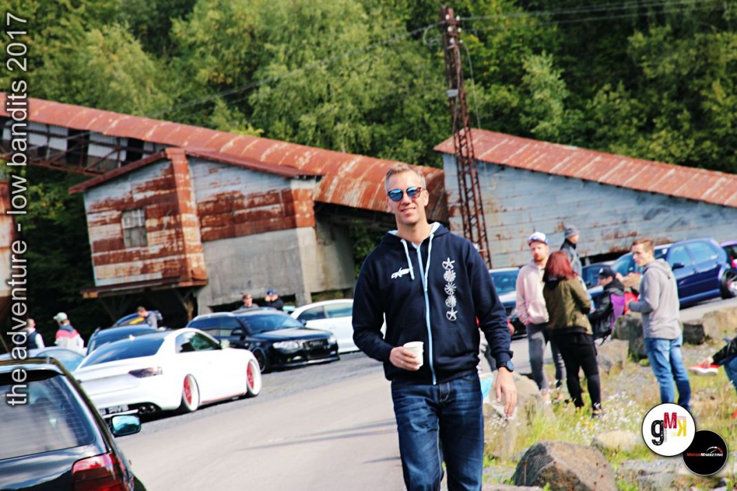 Teilnehmer in dunkler Jacke mit Sonnenbrille vor geparkten Youngtimern und Oldtimern in einem Steinbruch mit Industriegebäude