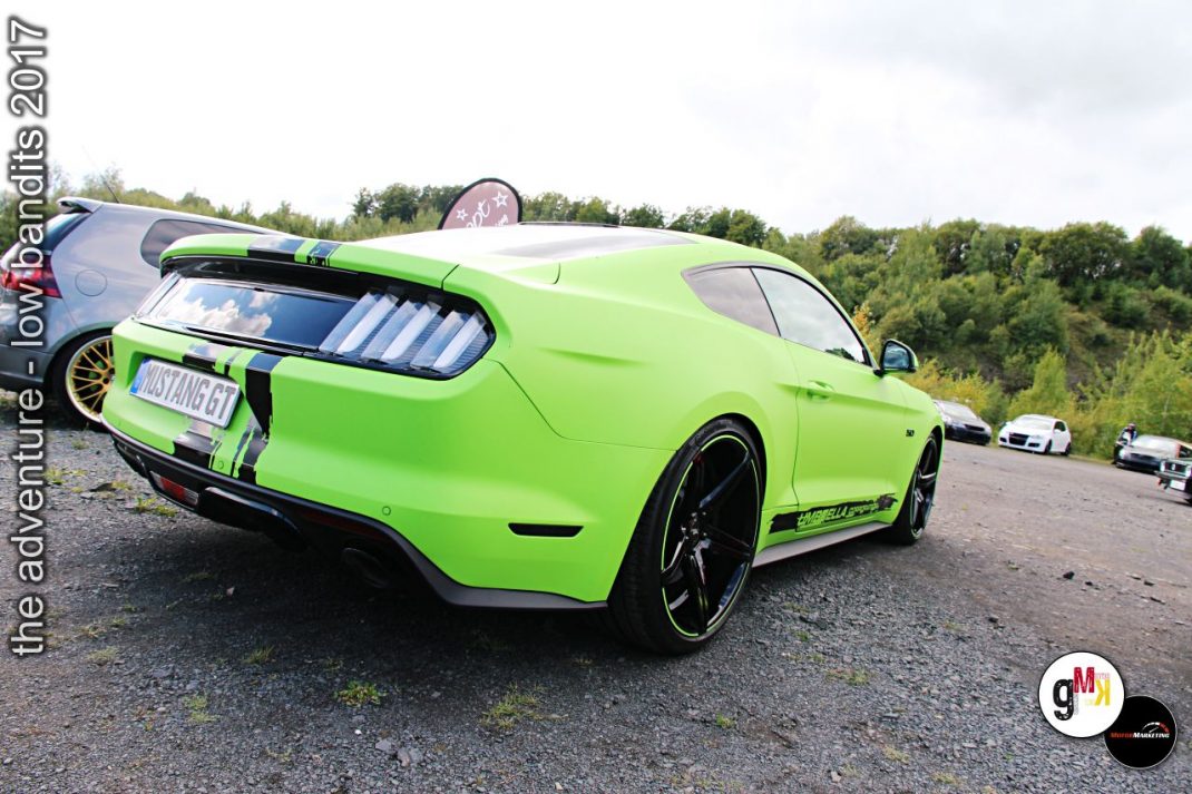 Neongrüner Muscle-Car von hinten auf einem Parkplatz mit anderen Fahrzeugen und Waldkulisse bei einem Tuning-Event
