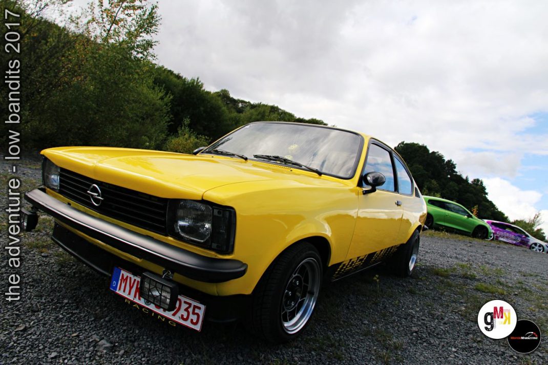 Gelber Opel-Oldtimer auf Schotterplatz bei Tuning-Treffen, weitere Youngtimer im Hintergrund unter bewölktem Himmel