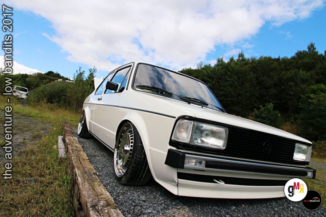 Weißer Youngtimer-Kompaktwagen mit getuntem Look parkt auf Schotterweg an Waldrand während Tuning-Event