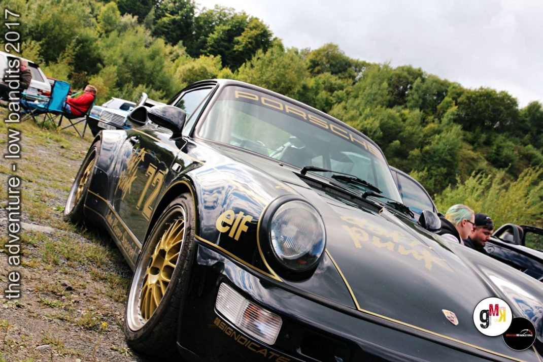 Schwarzer Porsche-Sportwagen mit goldenen Felgen und Elf-Schriftzug am Straßenrand, Teilnehmer mit Campingausrüstung im Hintergrund