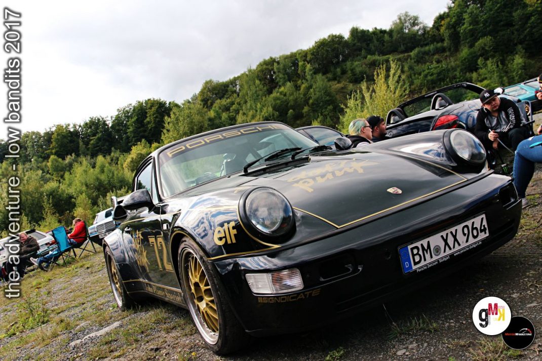 Schwarzer Porsche mit goldenen Felgen und Elf-Lackierung steht mit anderen Oldtimern an bewaldeter Strecke bei Rallye