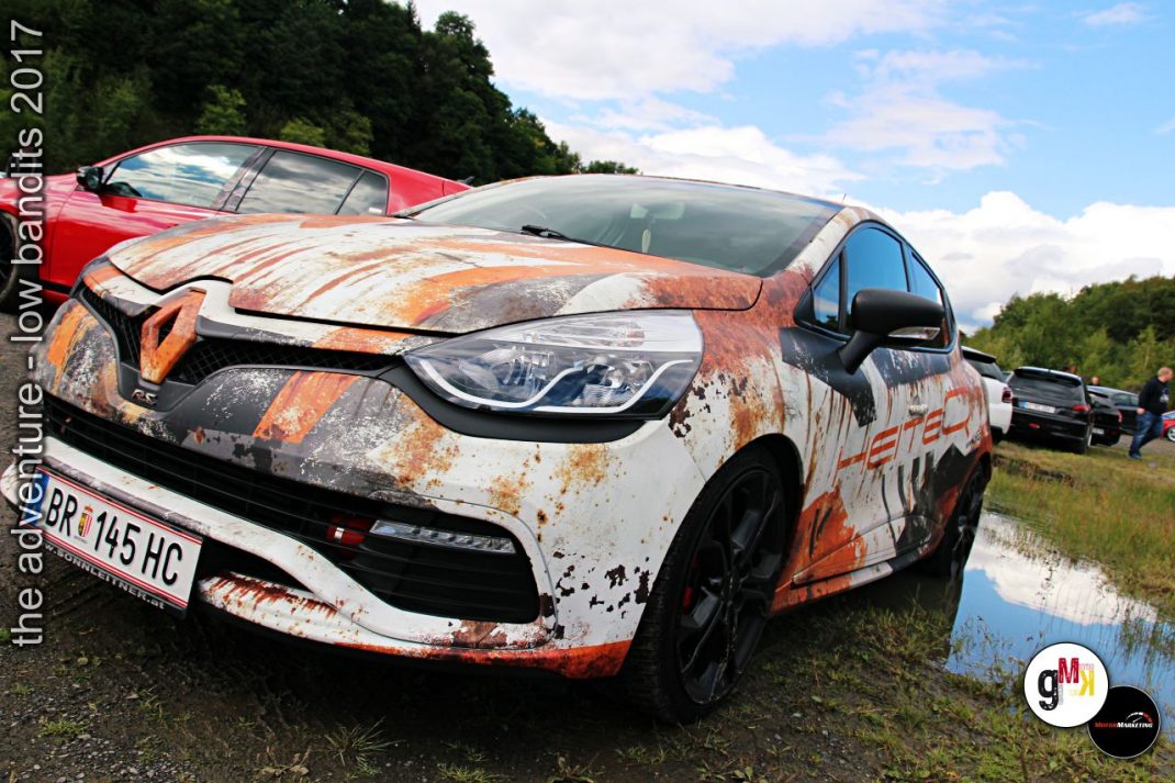 Youngtimer-Kompaktwagen mit orange-weißer Kunstlack-Folierung und Matsch-Effekt bei Adventure-Rallyefahrt, dahinter rotes Cabriolet