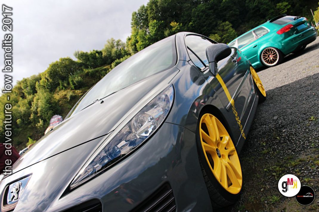 Silberner Sportwagen mit gelben Felgen und Abschleppseilen auf Parkplatz, im Hintergrund grünes Cabrio-Coupé bei Tuning-Event
