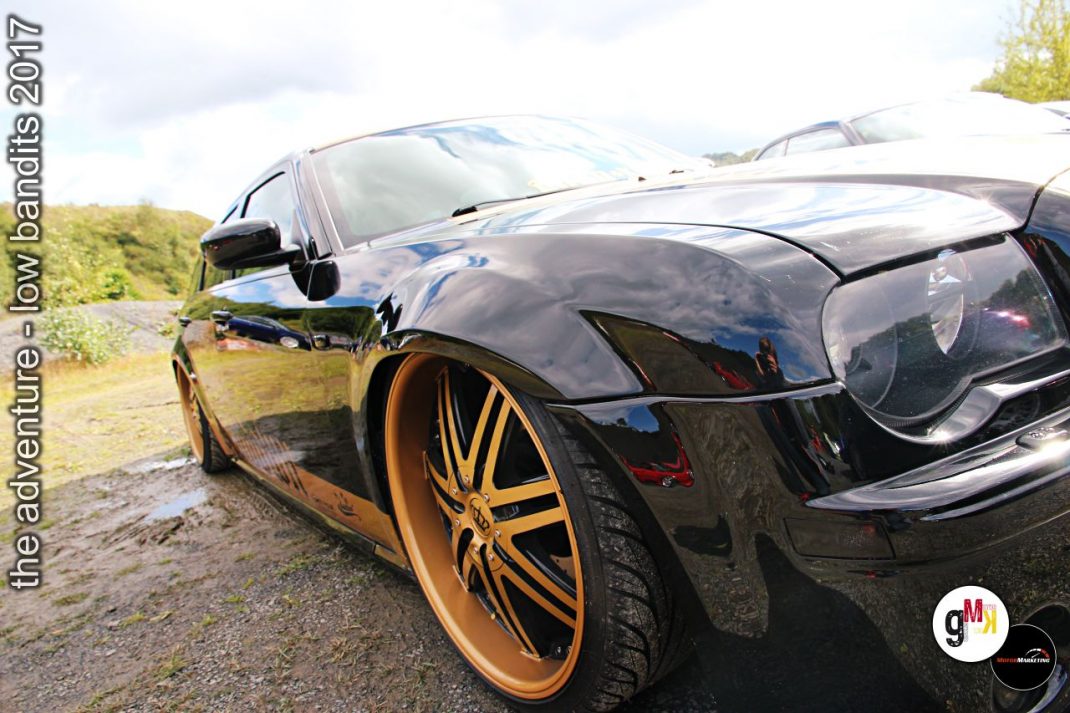 Schwarzer Sportwagen mit goldenen Leichtmetallrädern auf Treffen-Parkplatz, weitere Fahrzeuge im Hintergrund