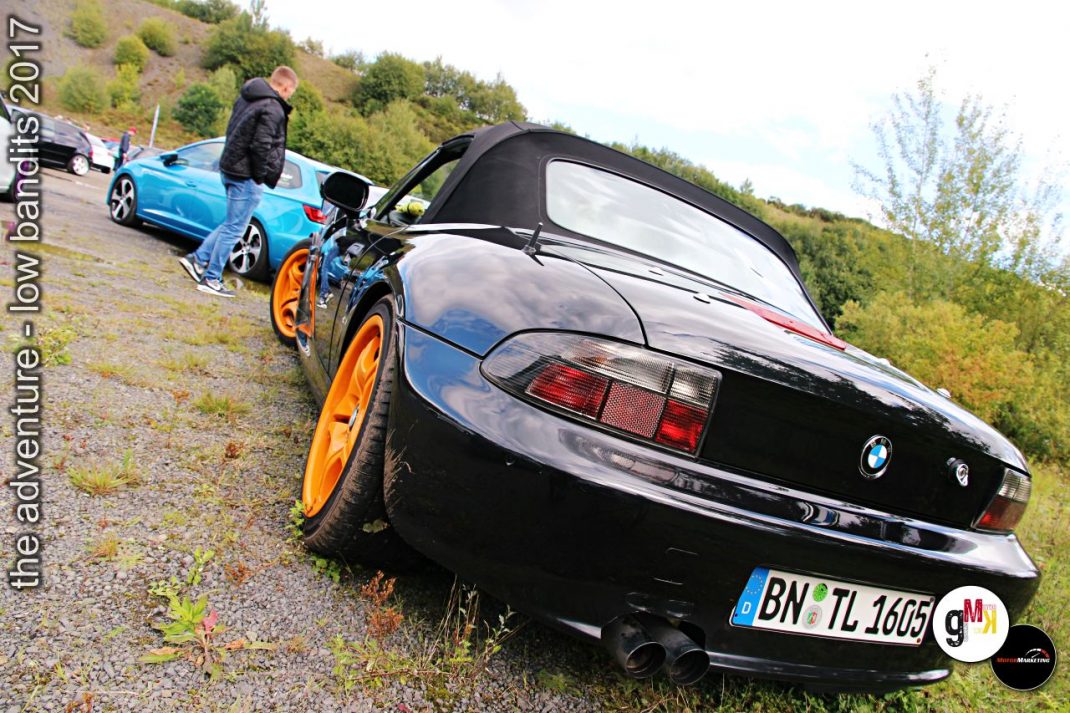 Schwarzes BMW-Cabrio mit orangefarbenen Felgen und blauer Youngtimer-Limousine auf Kiesplatz vor bewaldeter Böschung, Teilnehmer bei Low Bandits 2017