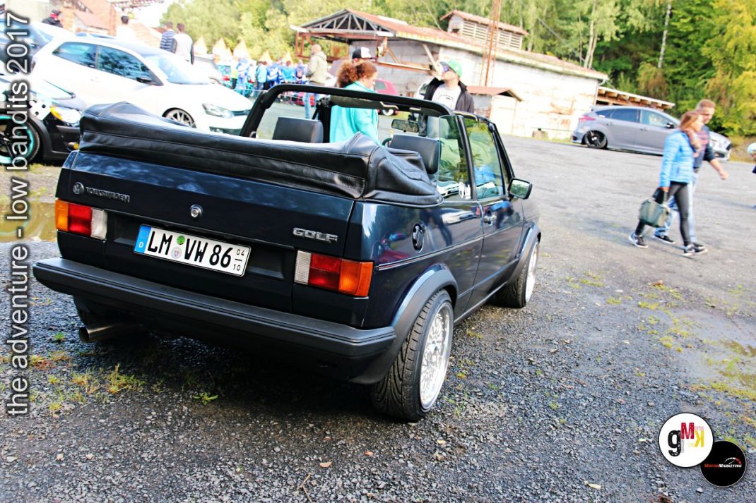 Schwarzer VW Golf Cabrio mit offenen Dach auf Parkplatz, Fahrer und Beifahrer sichtbar, Zuschauer und weitere Youngtimer im Hintergrund bei Tuning-Event