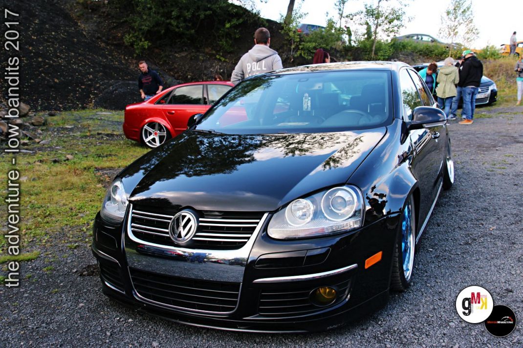 Schwarzer VW Jetta mit offener Motorhaube auf Veranstaltung, rotes Tuning-Auto im Hintergrund, Besucher auf Gelände
