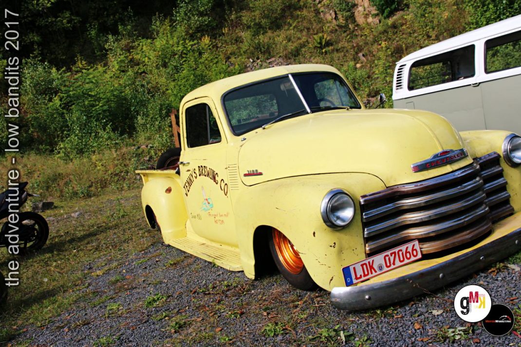 Gelber Oldtimer-Pickup mit Custom-Tuning und Vintage-Schriftzug parkt neben weißem Fahrzeug an Kiesplatz mit Waldkulisse bei der Low Bandits Veranstaltung 2017