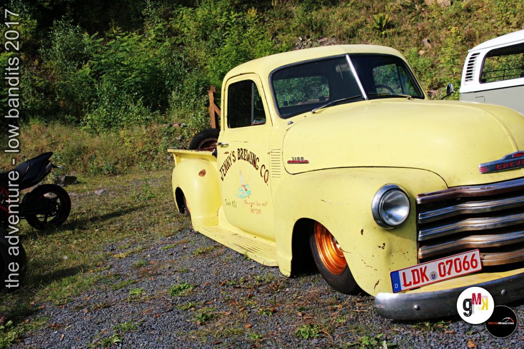 Gelber Oldtimer-Pickup mit Beschriftung steht auf Kiesplatz neben Vegetation, weitere Fahrzeuge im Hintergrund