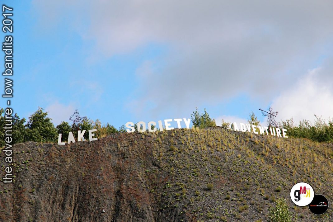 Großbuchstaben-Schriftzug „Lake Society" und „Lure" auf bewachsenem Steinbruch-Hang, Veranstaltungslogo unten rechts