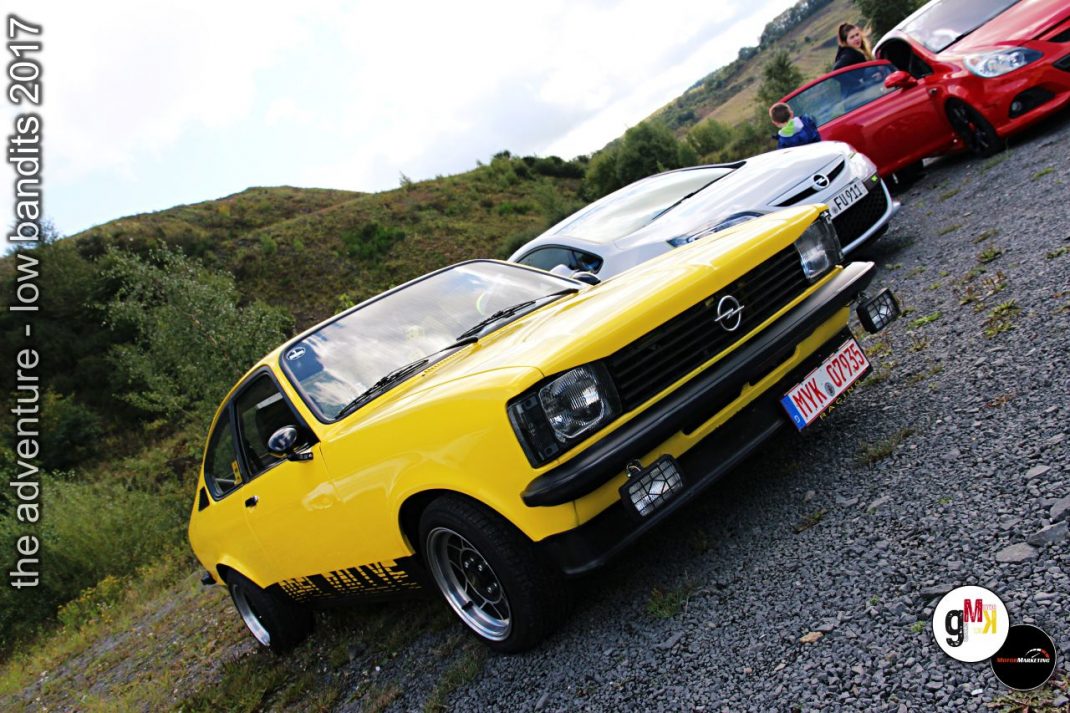 Gelber Opel-Oldtimer und weitere Youngtimer in Reihe auf Kiesplatz vor grüner Hügellandschaft beim Low Bandits Event