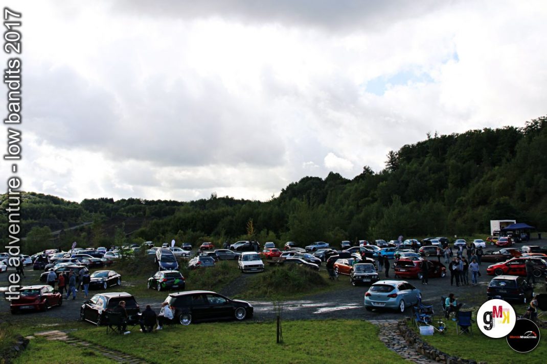 Parkplatz mit Oldtimern und Youngtimern auf Wiese vor bewaldeter Hügellandschaft, Besucher zwischen den Fahrzeugen bei Tuning-Veranstaltung